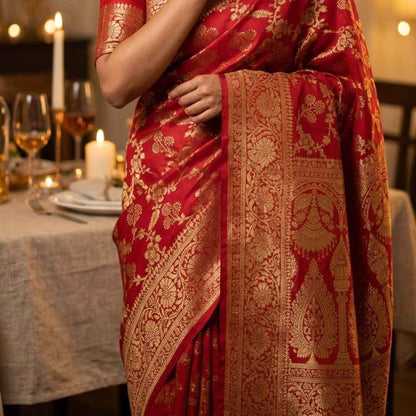 Woman in a red saree with gold patterns standing in a decorated indoor setting.